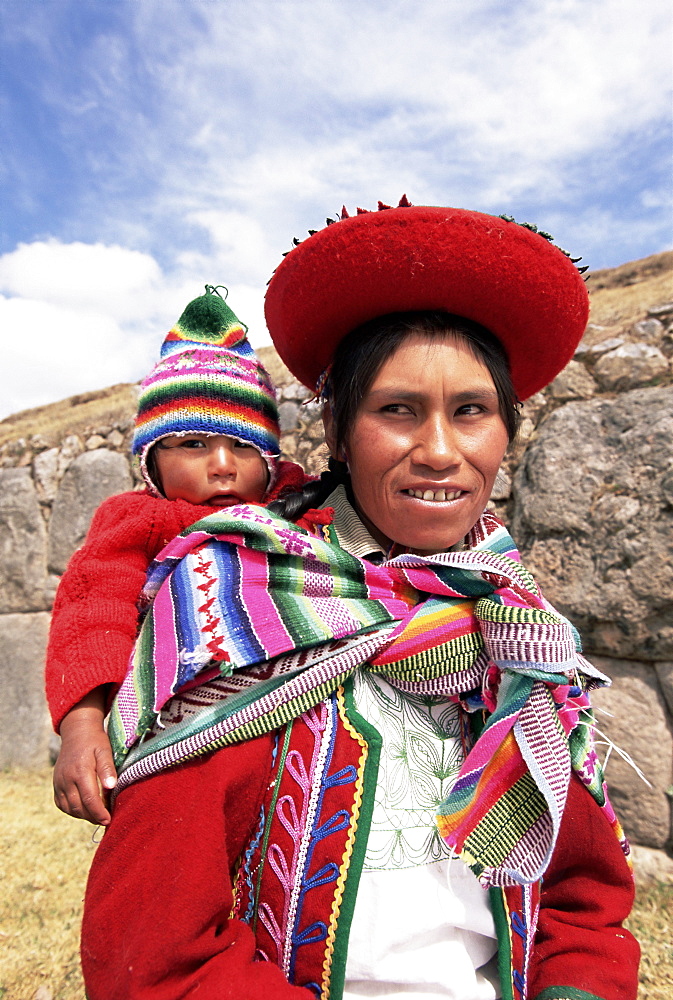 Portrait of a local woman in traditional dress, carrying her baby on her back, in front of Inca ruins, near Cuzco, Peru, South America