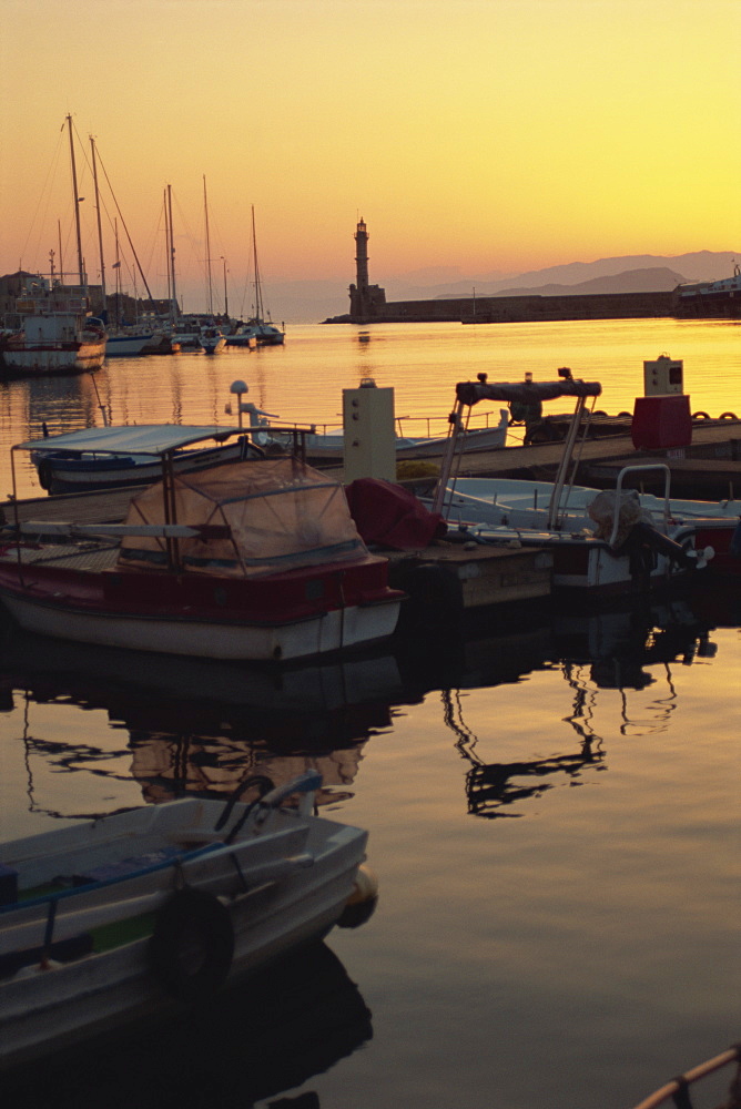 Dusk light over moored boats in the harbour and the lighthouse silhouetted on the horizon, Chania, Crete, Greece, Europe