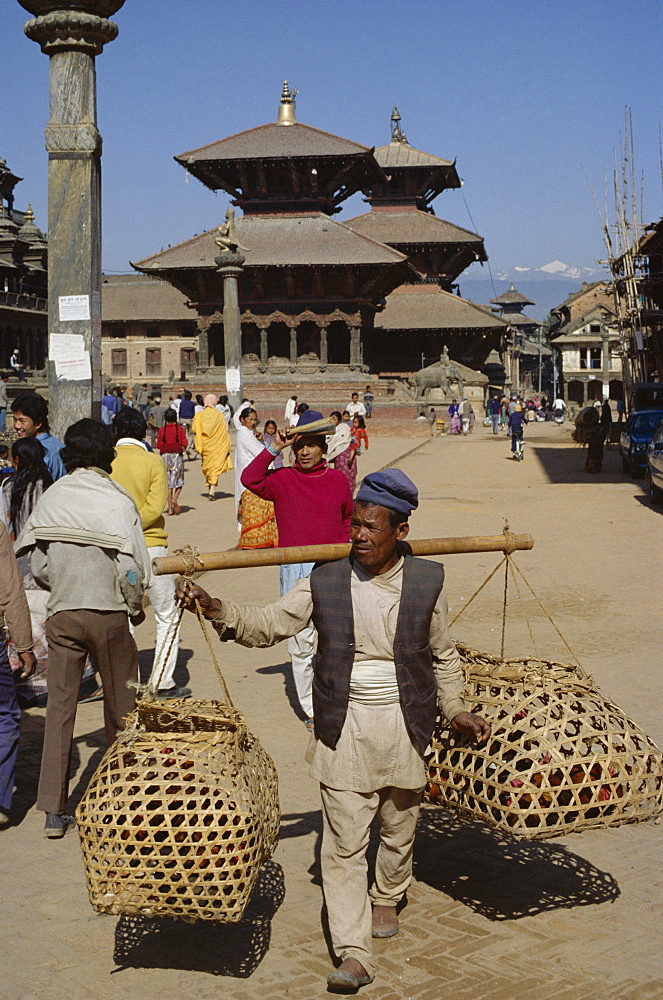 Nepali man carrying produce in wicker baskets, with Garuda statue, Bishwan Nath Mandir and Bhimsen Mandir temples dating from 17th century behind, Durbar Square, Patan, Kathmandu, Nepal, Asia