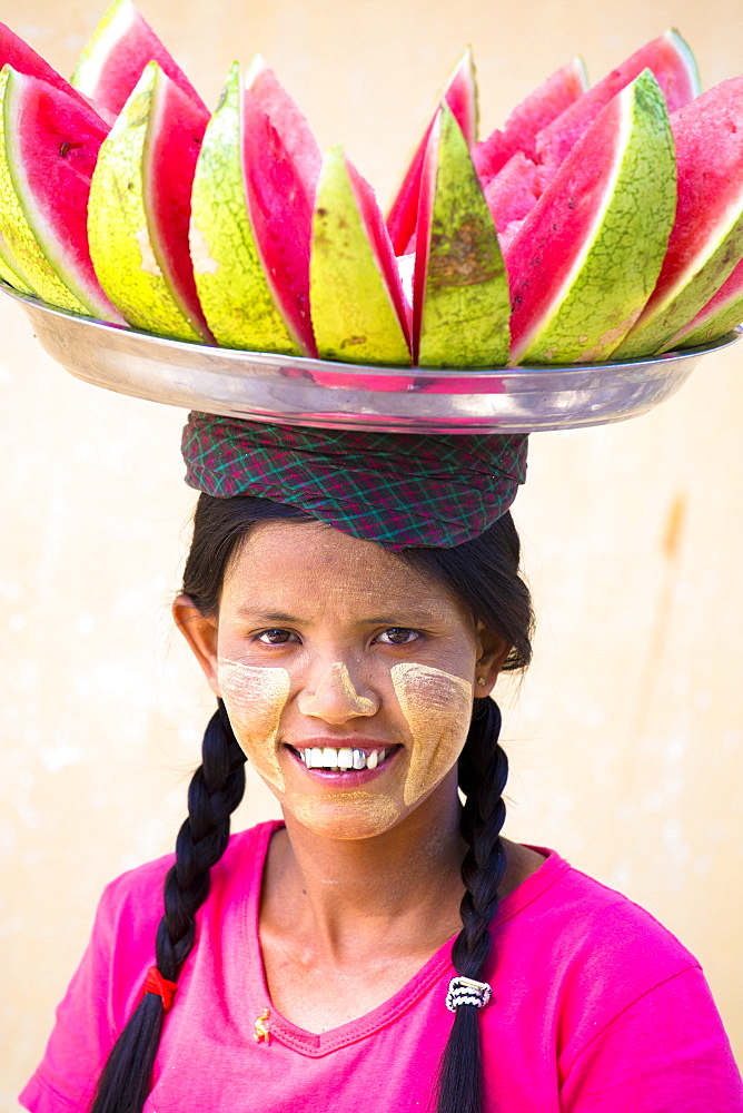 Local woman with Thanakha traditional face painting, carrying a tray of sliced watermelon on her head, Shwezigon Paya, Nyaung U, Myanmar (Burma), Asia