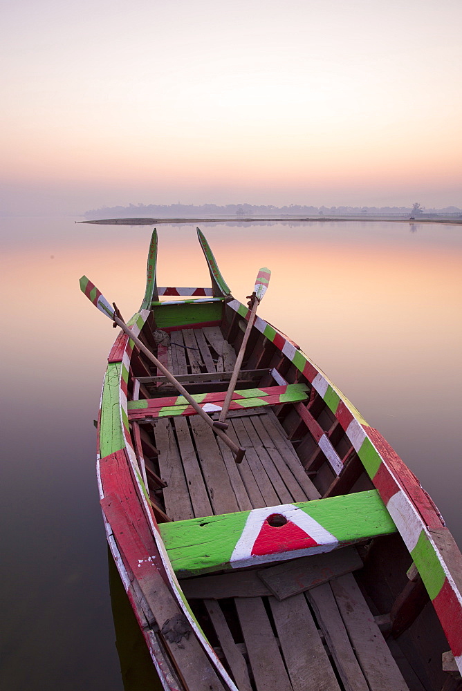 Traditional rowing boat moored on the edge of flat calm Taungthaman Lake at dawn with the colours of the sky reflecting in the calm water, close to the famous U Bein teak bridge, near Mandalay, Myanmar (Burma), Asia