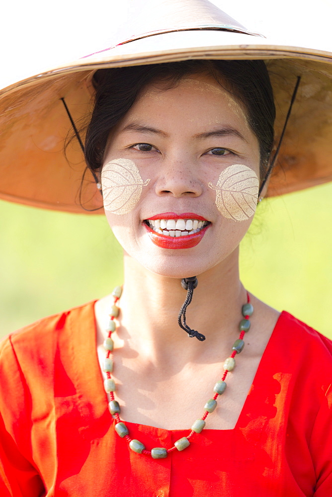 Portrait of local woman wearing traditional bamboo hat, red dress and Thanaka face painting in the shape of leaves on her cheeks, near Mandalay, Myanmar (Burnma), Asia