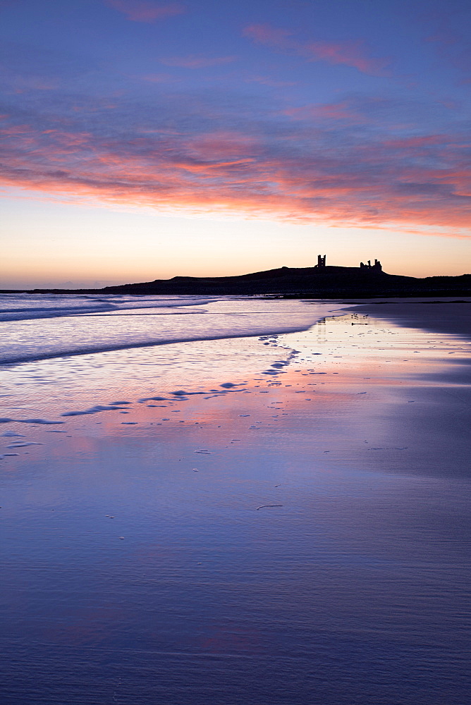 Looking across Embleton Bay at sunrise towards the silhouetted ruins of Dunstanburgh Castle in the distance and the vivid colours in the sky reflecting in the sea and wet sand, Embleton, near Alnwick, Northumberland, England, United Kingdom