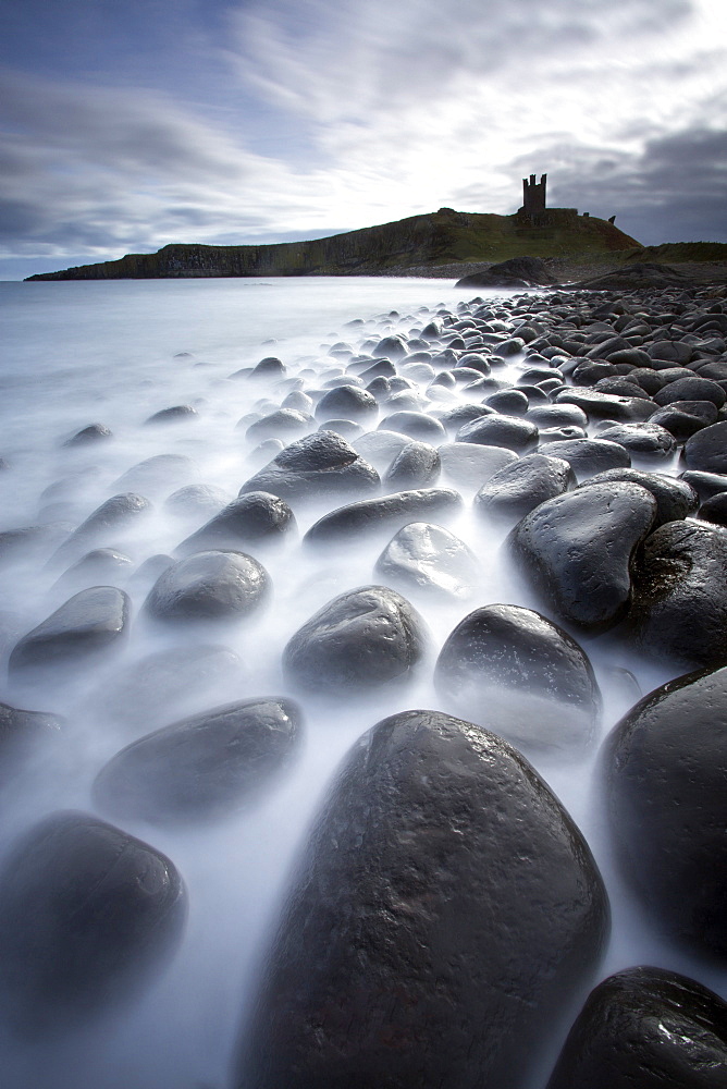 Long exposure to record motion in the sea as it washes around black basalt boulders on a beach at Embleton Bay with the silhouette of Dunstanburgh Castle in the distance, Embleton Bay, near Alnwick, Northumberland, England, United Kingdom, Europe