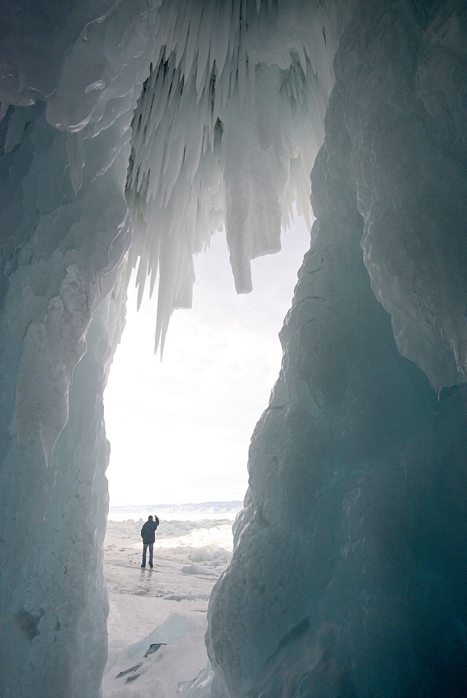 Tourist framed in the frozen mouth of ice cave, with icicles formed on roof of caves at Olkhon Island as the waves freeze in winter, Lake Baikal, Irkutsk Oblast, Siberia, Russia, Eurasia