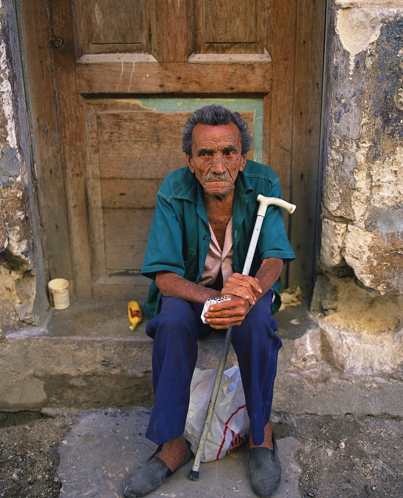 Portrait of an elderly man with a stick sitting on a doorstep selling cigarettes in Old Havana, Cuba, West Indies, Central America