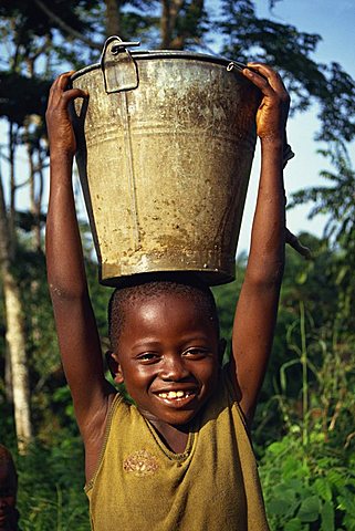 Head and shoulders portrait of a young African child carrying a bucket of water on his head, smiling and looking at the camera, Liberia, West Africa, Africa