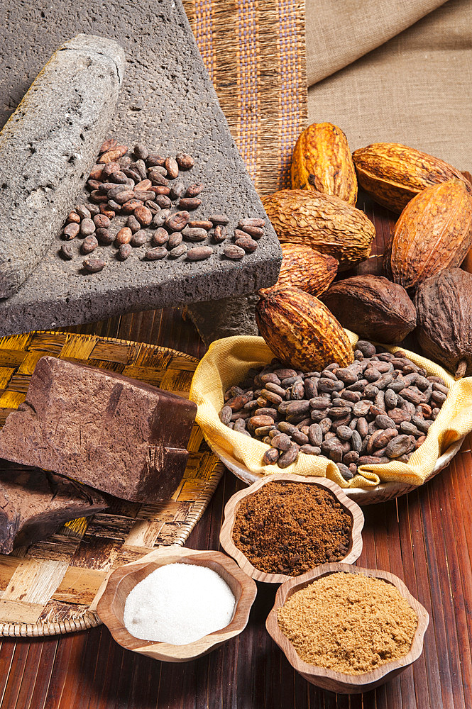 A still life with an ancient stone Metate (A flat stone with a slightly concave surface, used with another stone for grinding cocoa seeds) and raw cocoa, chocolate, cocoa beans, cabosse, assorted spices and different types of sugar.