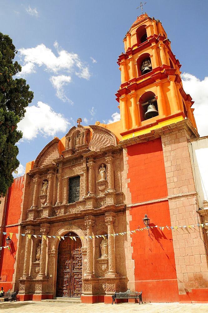 Mexico, Bajio, San Miguel de Allende, Exterior facade and bell tower of orange and yellow painted church building with flags hanging across doorway.