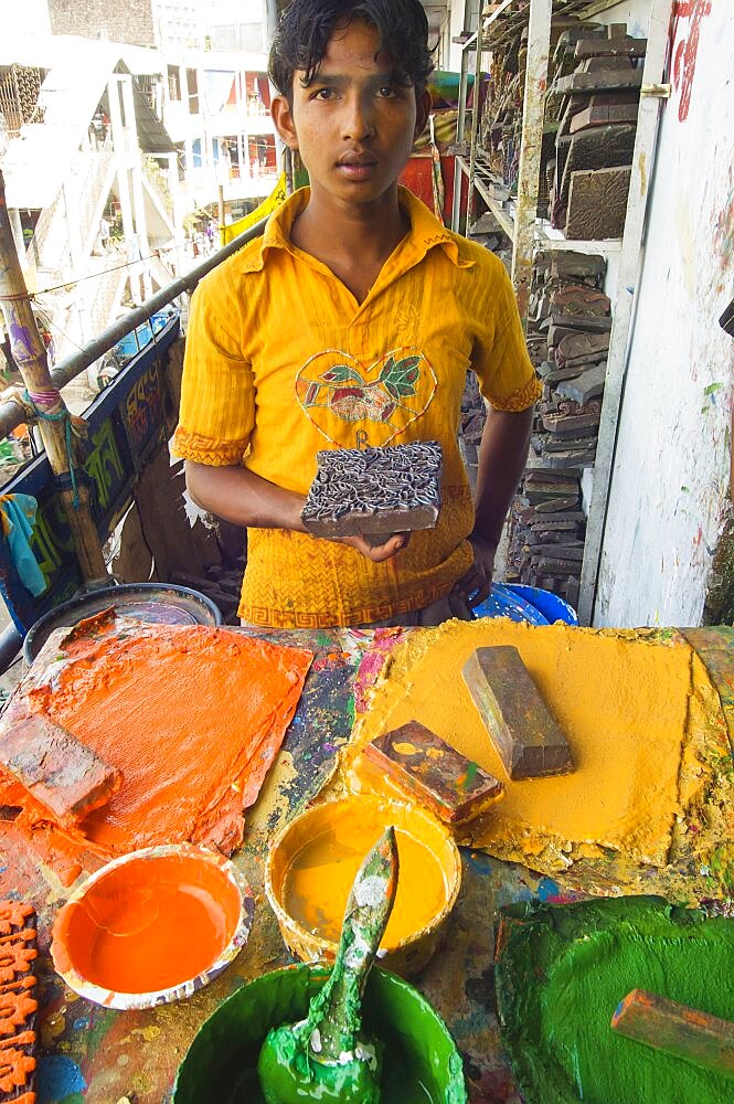 Bangladesh, Dhaka, Young man holding a printing block used to print on cotton fabrics in New Market with pallettes of colours in front of him.