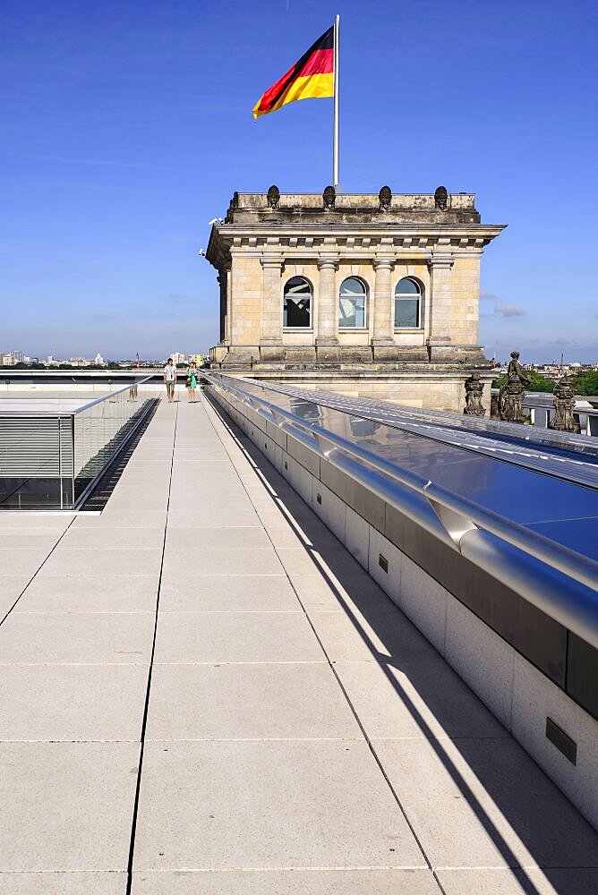 Germany, Berlin, German flag fluttering on a corner tower of the Reichstag building as seen from the rooftop terrace with two tourists strolling.