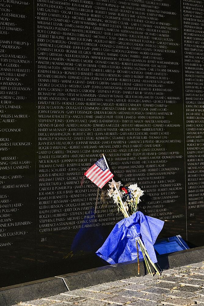 USA, Washington DC, National Mall, Vietnam Veterans Memorial, A section of The Memorial Wall with the names of those killed or missing in action during the Vietnam War, Bunch of flowers and American flag resting against the wall.