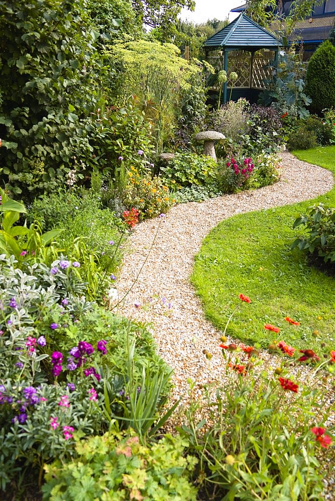 English cottage garden, winding shingle path leading to a gazebo between grass lawn and flowerbed of mixed plant varieties.