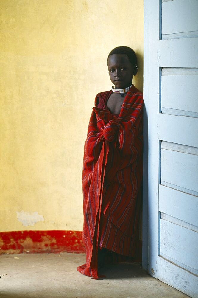 TANZANIA  Ngorongoro Masai child standing between yellow wall and blue door.  The Masai have been expelled from the Ngorongoro Crater area to make way for tourists and wildlife.