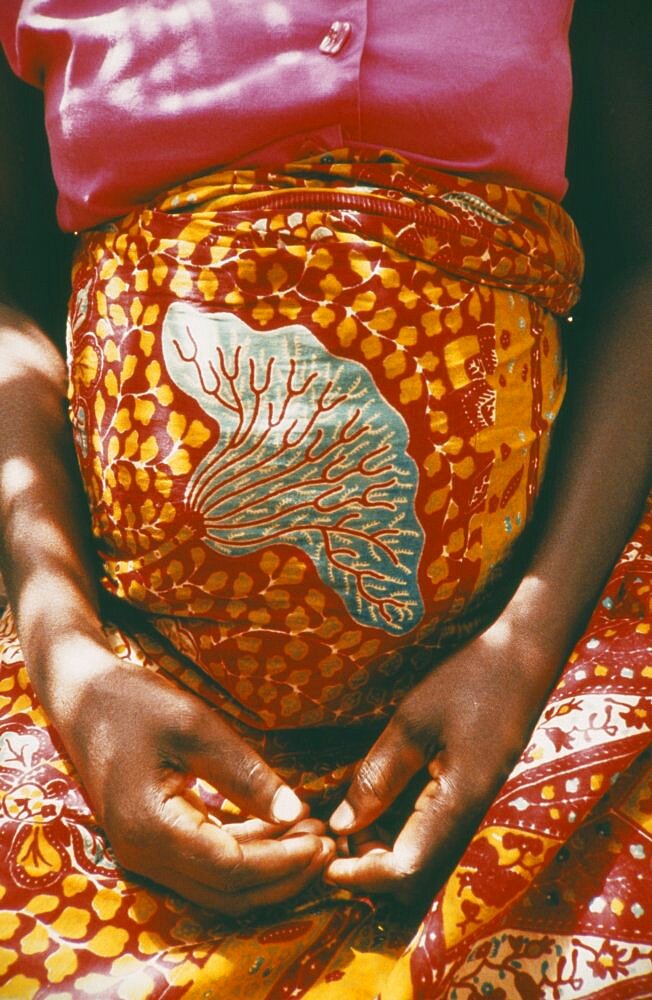 GHANA  Volta Region Cropped shot of woman thirty six weeks pregnant wearing brightly coloured sarong and with her hands folded in her lap beneath her belly. Colored Leis