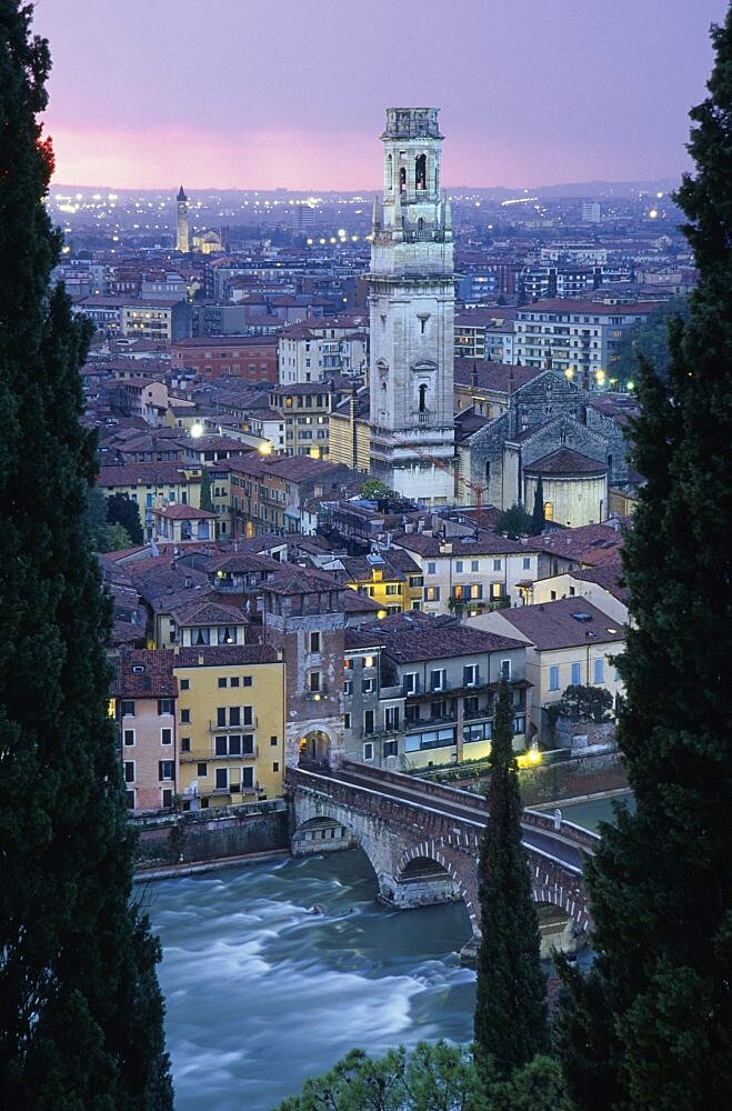 ITALY Veneto Verona Cityscape at sunset showing tiled rooftops  Duomo bell tower and bridge over the Adige River part framed by tall coniferous trees.  Pale pink/purple sky and city lights.