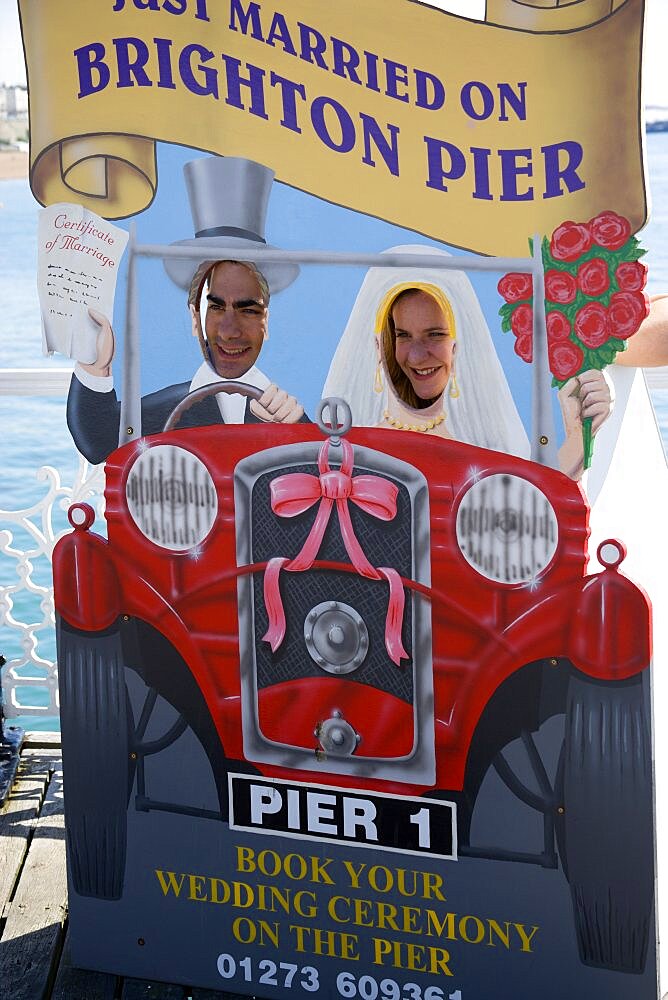 England, East Sussex, Brighton, Man and woman with their faces in amusement cut-out of wedding car that says Just Married On Brighton Pier which also serves as a bookings advertisement for marriage ceremonies on the Pier itself.