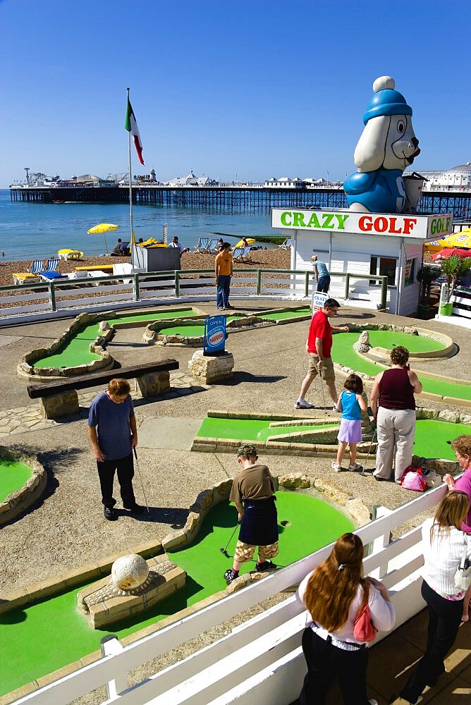 England, East Sussex, Brighton, Adults and children playing miniature golf on a Crazy Golf course on the promenade with Brighton Pier and the pebble shingle beach beyond.