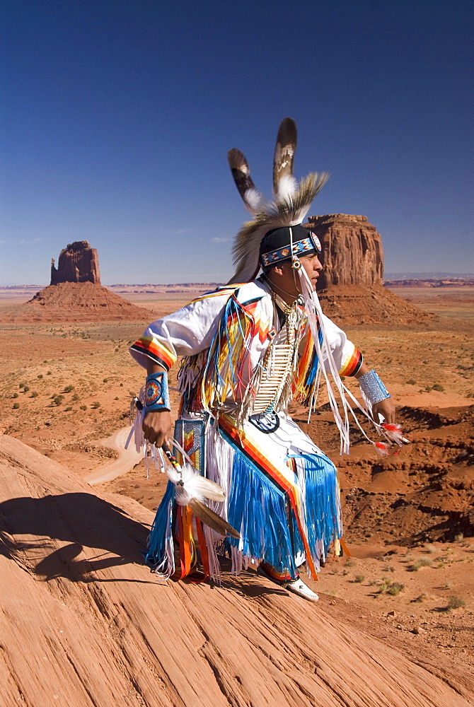 Navajo man dressed in traditional costume dancing, with Merrick Butte on the right and East Mitten Butte on the left in the background, Monument Valley Navajo Tribal Park,  Utah, United States of America, North America