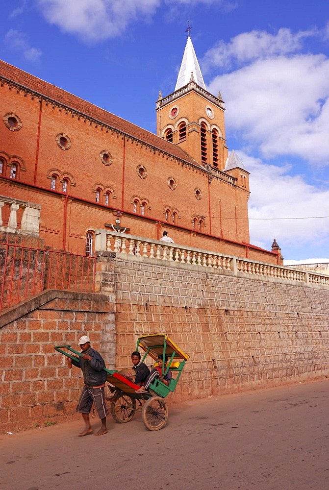 Man transporting passenger in his rickshaw on foot beside the red brick cathedral in Ambositra, Madagascar, Africa