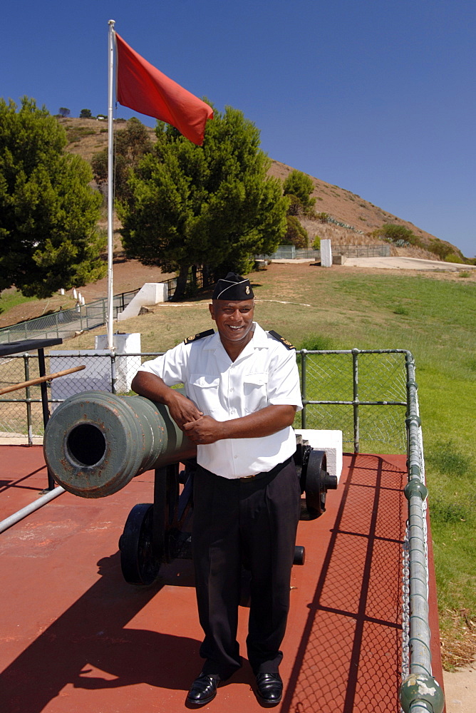 Chief Petty Officer Dudley Malgas of the South African Navy posing alongside the noon gun cannon in Cape Town. CPO Malgas has been in charge of firing the canon since 1995.

The daily noon gun is Cape Town’s oldest living tradition and the two cannons used are the oldest guns in daily use in the world. They have marked the midday hour in the mother city in this distinctive, albeit noisy manner since early 1806. The cannons were cast in Britain in 1794 and still bear the royal crest of King George the third. The firing of the cannon was originally to give ships in the bay a means of re-setting their clocks accurately.