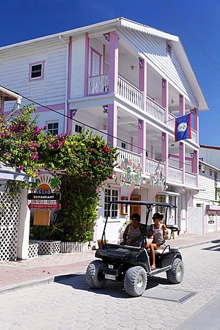 Two women in a golf cart driving past a wooden house painted in Caribbean colors, San Pedro, Ambergris Cay Island, Belize, Central America, Caribbean