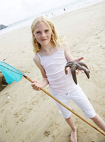 Blonde girl standing on the beach, smiling, holding a fishing net and a starfish