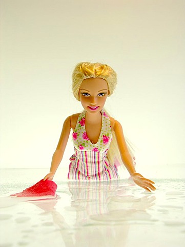 A blond Barbie doll kneels in a flower dress before white background on a white soil with finery water and a cloth to clean the floor