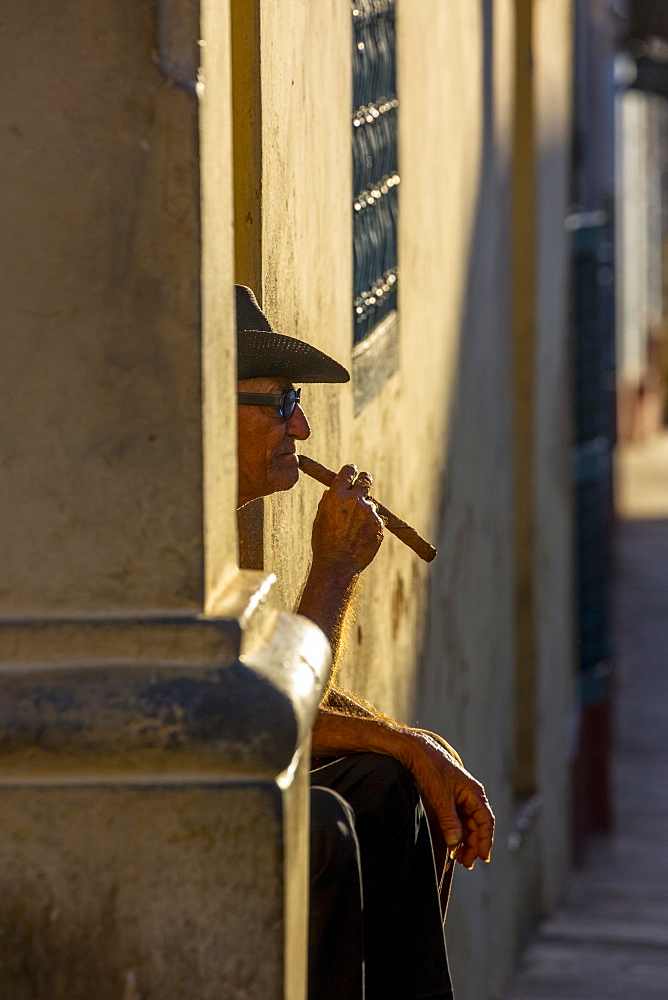 An elderly Cuban sitting on a doorstep in the evening light, smoking a cigar, Trinidad, Sancti Spiritus Province, Cuba, Central America