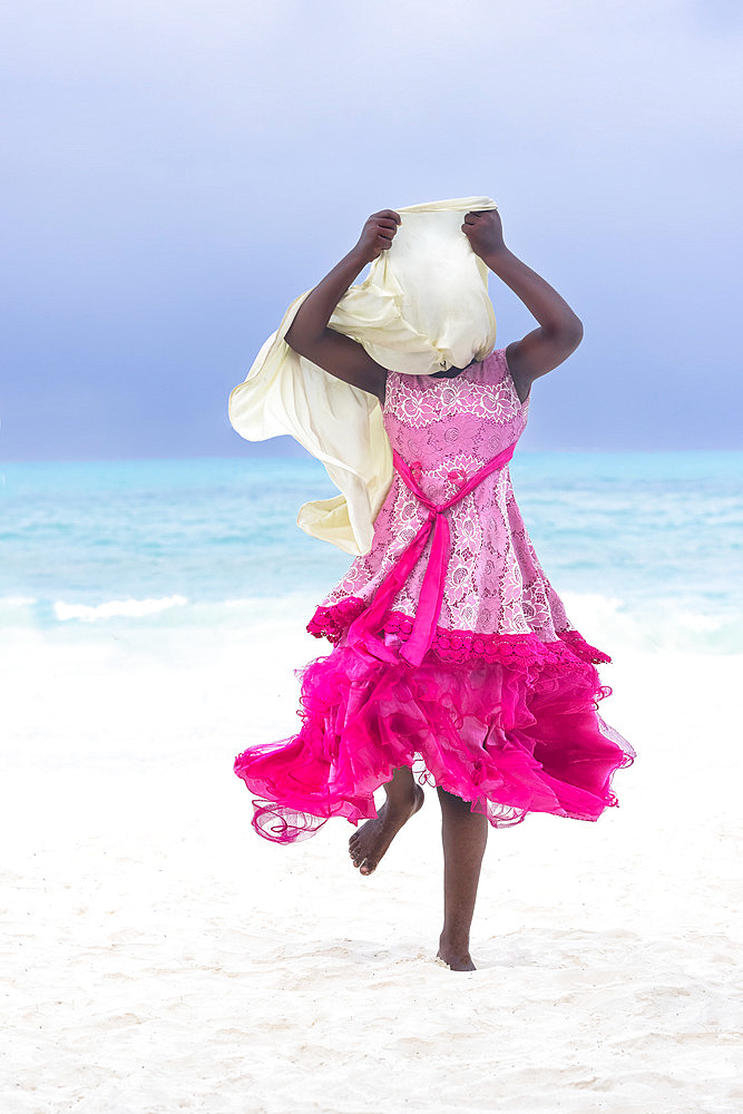 Young girl dressed in a pink fuchsia dress with ruffles and veil, white hijab. In the late afternoon, children from a small village come to play by the sea, a beach of white sand and turquoise sea on the east coast of Zanzibar, Tanzania.