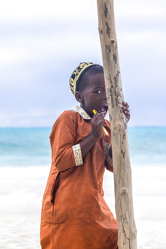 Portrait of a little boy wearing an orange abaya and a black and gold kofia, hiding behind a wooden pole. In the late afternoon, children from a small village come to play on the white sand beach and turquoise sea on the east coast of Zanzibar, Tanzania..,