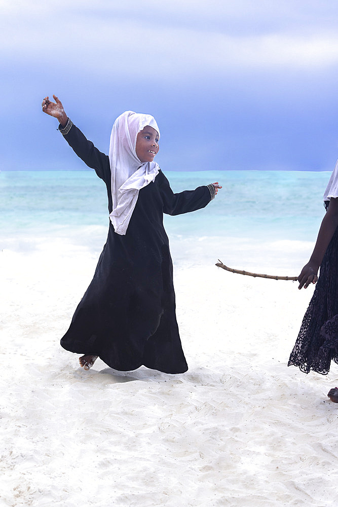 Two young girls dressed in white veils and black abaya, the older one threatens the younger one with a stick while the latter is dancing in defiance. In the late afternoon, children from a small village come to play on the white sand beach and turquoise sea on the east coast of Zanzibar, Tanzania.