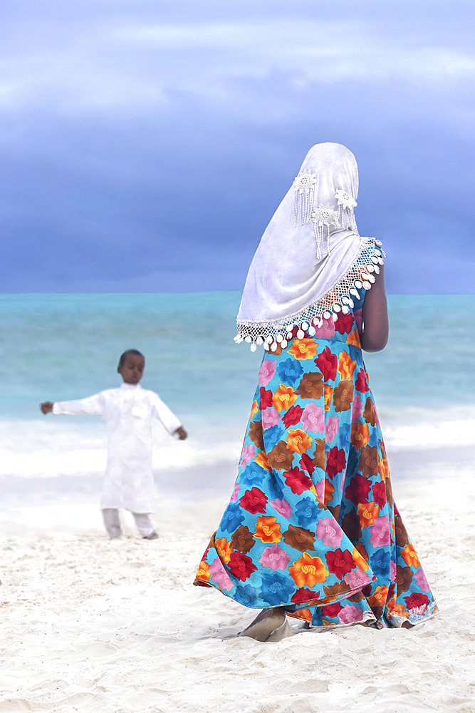 Young girl dressed in a blue dress with orange, red and brown floral motifs and a white hijab veil. In the late afternoon, children from a small village come to play on the white sand beach and turquoise sea on the east coast of Zanzibar, Tanzania.