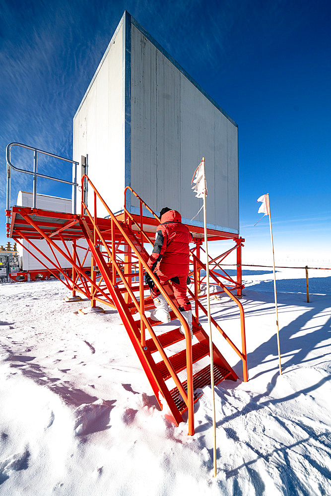 A technician goes into a raised square building on an orange metal structure. An Italian technician specialising in radio and telecommunications goes into the shelter that houses the VSAT antenna, the device that connects the station to the Internet by satellite. This is the station's only link with the outside world. Most of the buildings are elevated to prevent the formation of snowdrifts caused by wind-blown snow. Concordia Antarctic Research Station, Dome C plateau, East Antarctica.