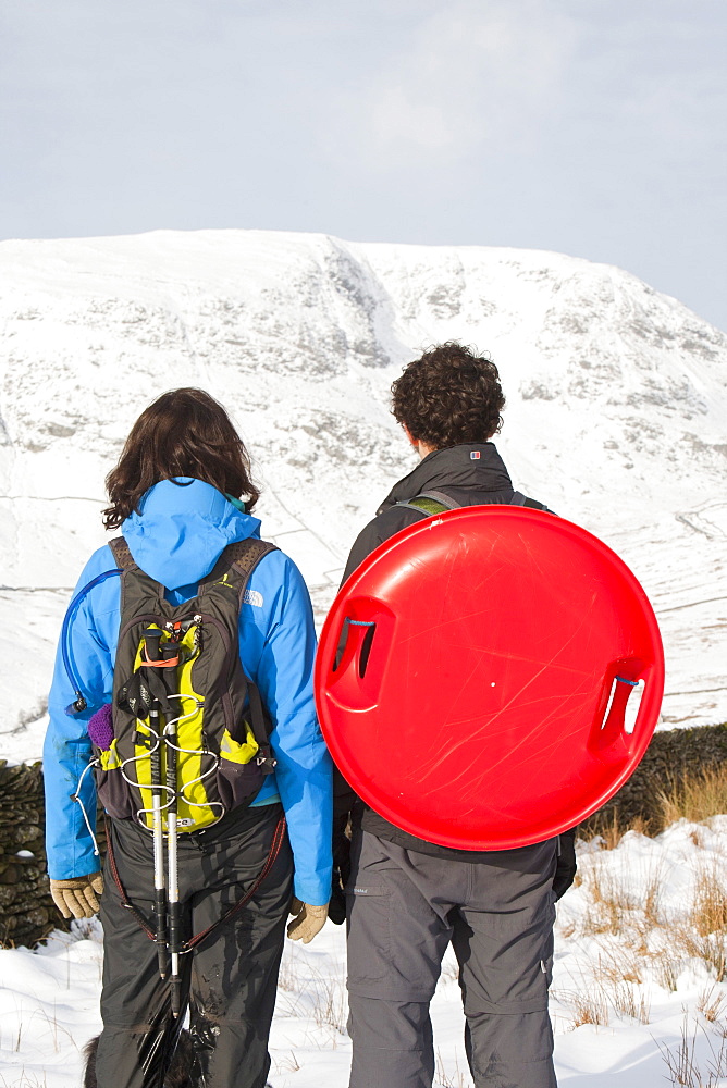 A couple with a plastic sledge in the snow on Wansfell above Ambleside in the Lake District, looking towards Red Screes, Cumbria, England, United Kingdom, Europe