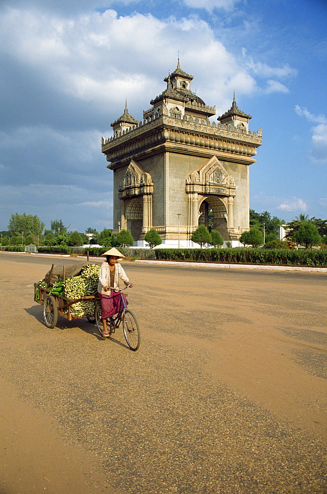 Woman on a bicycle with a trailer rides past the Anousavari monument in Vientiane, Laos, Indochina, Southeast Asia, Asia