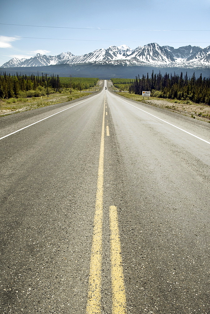 Long straight road leading to dramatic mountain range, snow capped mountains with forrest either side.  Clear blue sky in Sunny Alaska Interior. The Interior, Alaska, USA