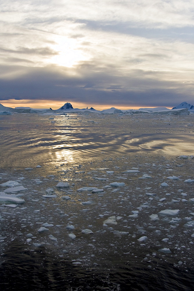 Sunset on snow capped mountains and icebergs surrounded in brash ice in Crystal Sound near the Antarctic Circle on the west side of the Antarctic Peninsula.