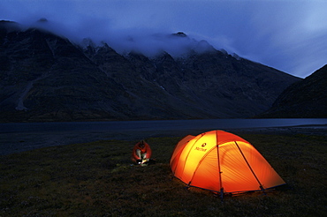 A man with a headlamp in front of a glowing tent on the shores of Russvatnet in Jotunheimen National Park, Norway.