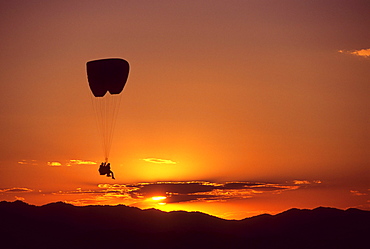 A tandem paraglider pilot and passenger soar over Jackson Hole, Wyoming at sunset.