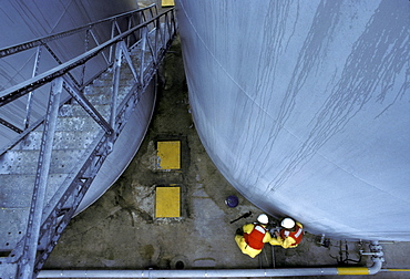 Two men working at the bottom of two cylinder shaped silos are visible from above a ladder at an oil field facility offshore.