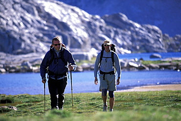 Two men backpacking in an alpine meadow with a lake in Eastern Sierra Nevada mountains, California.