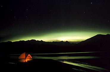 The aurora borealis (northrn lights) glows above a tent in the Knik River Valley. The Chugach Mountains are in the background.