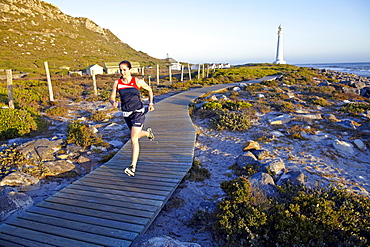 Susann Scheller running on a wooden boardwalk near the ocean and Kommetjie Lighthouse. South Africa.