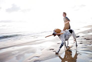 A young boy and a dog with a stick in his mouth   run along a quiet beach in Mexico.