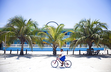 A little girl rides her bike on the sand in front of a colorful building and a row of palm trees.