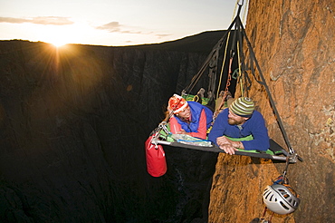 A man and woman on a portaledge at sunset while rock climbing a vertical face in Gunnison, Colorado.