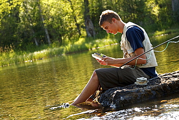 A teenage boy sits on a rock as he chooses a fly while fly fishing on the Swan River near Bigfork, Montana.