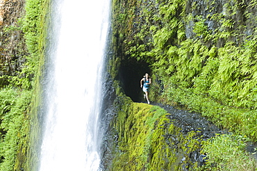 A woman running along trail blasted into a hill behind a waterfall on the Pacific Crest Trail, Oregon.