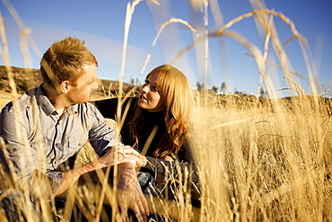 Couple sitting on the ground of an open field look at each other and smile.